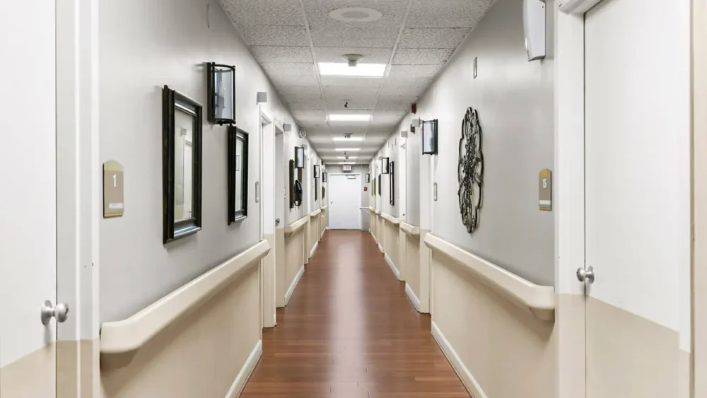 A well-lit, narrow hallway with wooden flooring, framed pictures on walls, and numbered doors on either side, leading to an exit