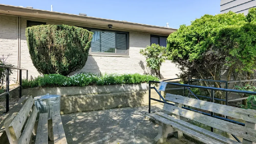 An outdoor area with two wooden benches, a concrete patio, and a ramp with a black handrail next to a brick building
