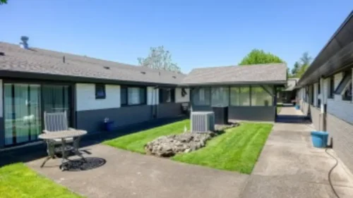 A courtyard with a grassy area and a patio with a table and chairs, between two buildings with dark roofs and siding