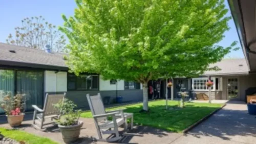 A courtyard area with a large tree, a path, and two white rocking chairs on a patio next to a building