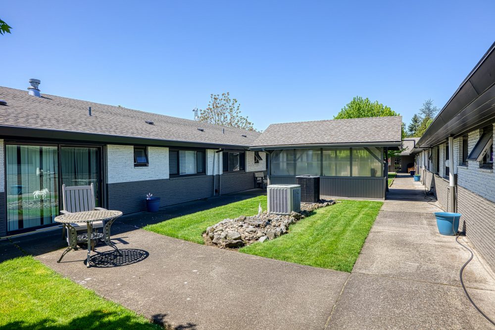 A courtyard with a grassy area and a patio with a table and chairs, between two buildings with dark roofs and siding
