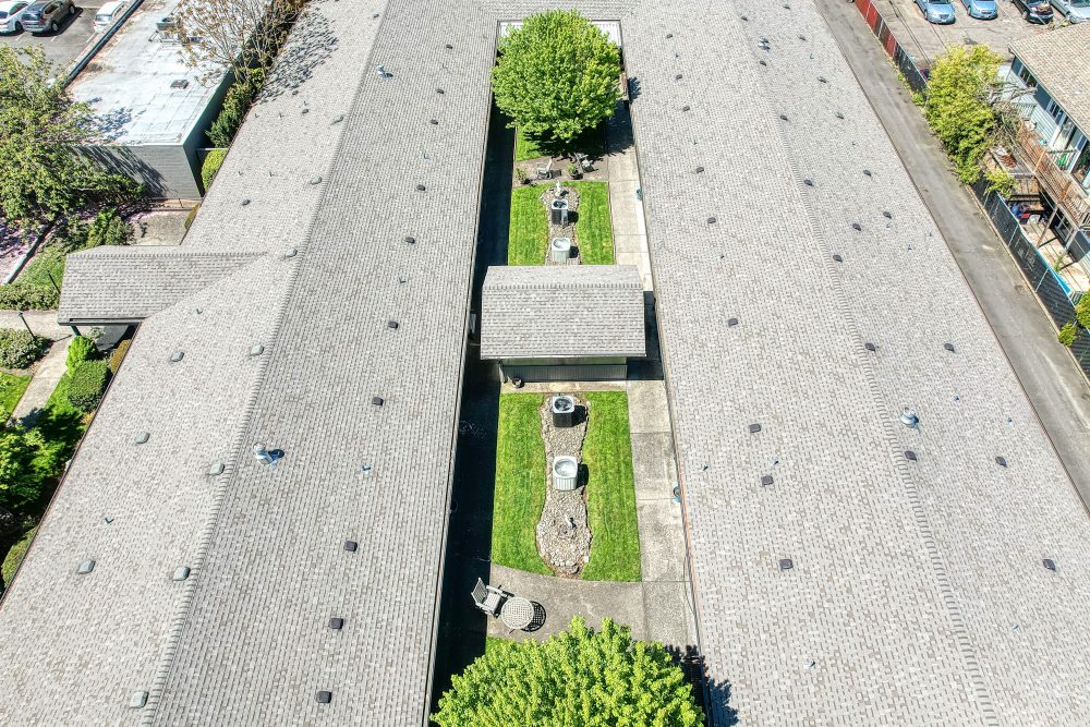 An aerial view of two rows of buildings with gray roofs, separated by a grassy courtyard with a tree and a fountain